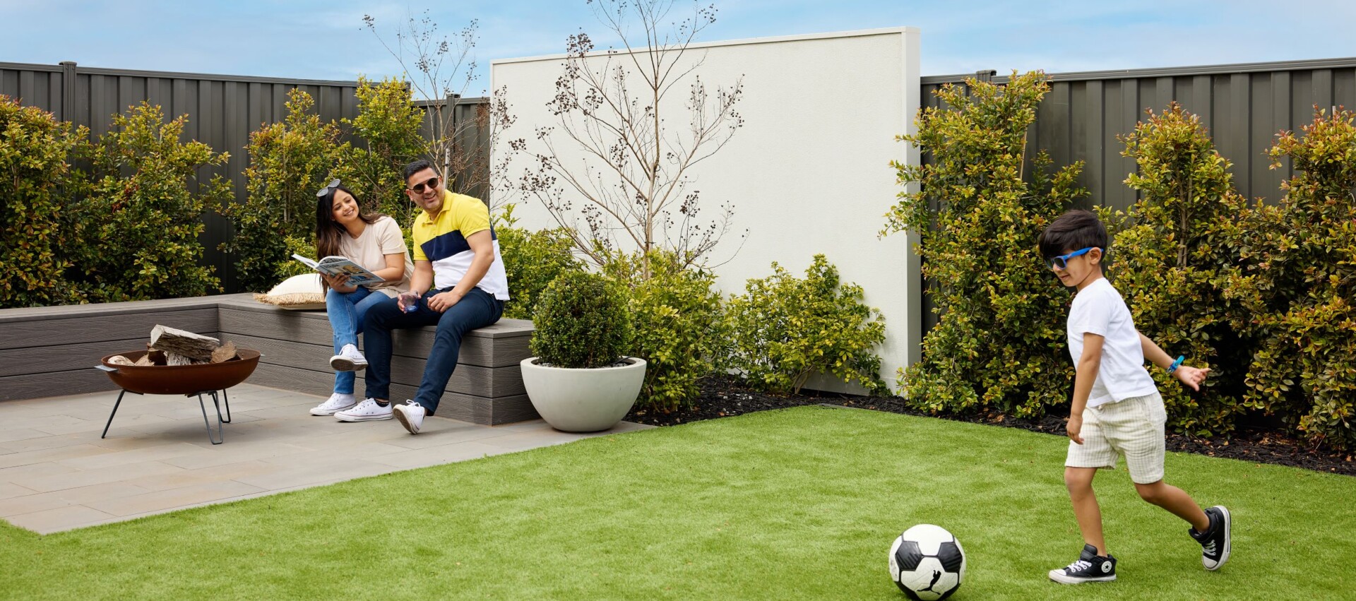 Young family enjoying backyard living in a modern Homebuyers Centre home, with kids playing soccer on the lawn and parents relaxing under a clear sky.