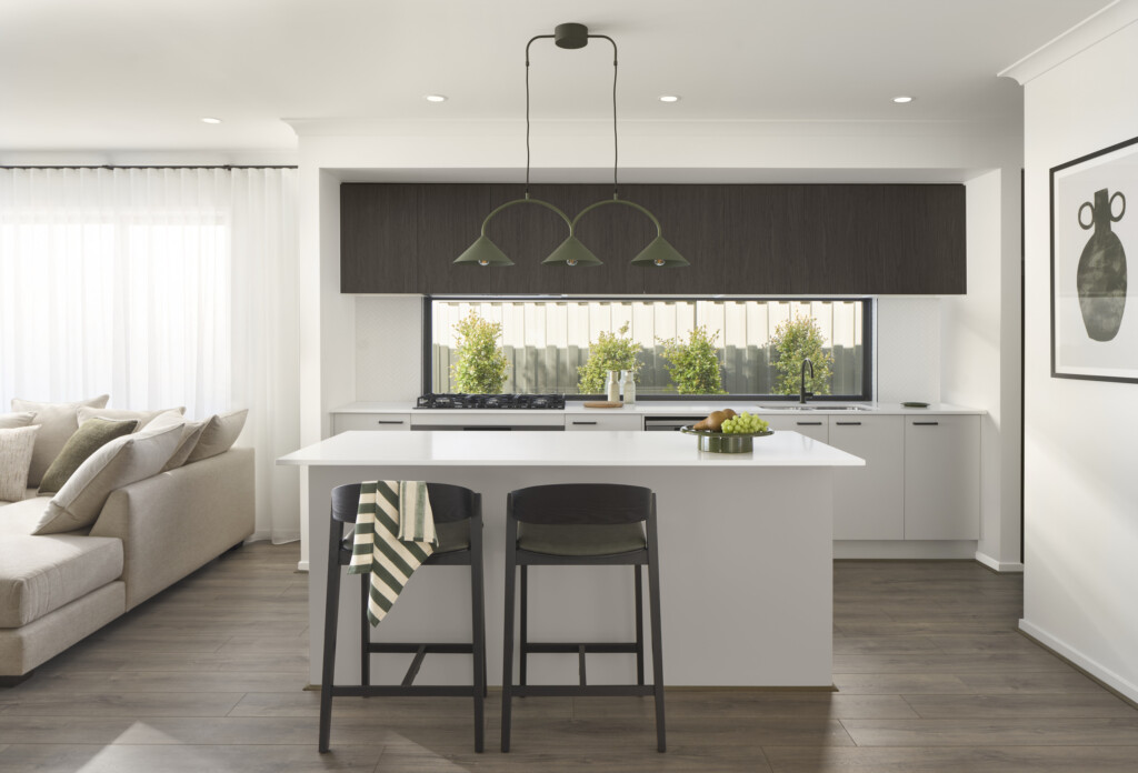 Modern kitchen in the Winton 25 display home at Lucas Estate Ballarat featuring white island bench, dark overhead cabinetry, window splashback, contemporary pendant lighting and open-plan layout.