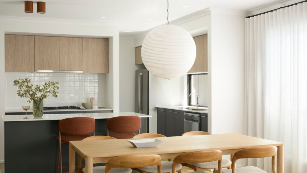 Open-plan dining and kitchen with a timber dining table and chairs, pendant light, breakfast bar with upholstered stools, light wood cabinetry, and soft natural light through sheer curtains.
