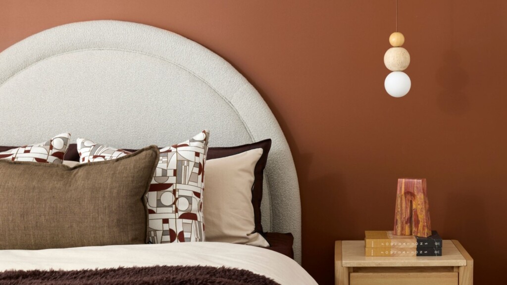 Modern bedroom interior featuring a curved upholstered bedhead, warm terracotta feature wall, styled cushions and timber bedside table with decorative lighting.